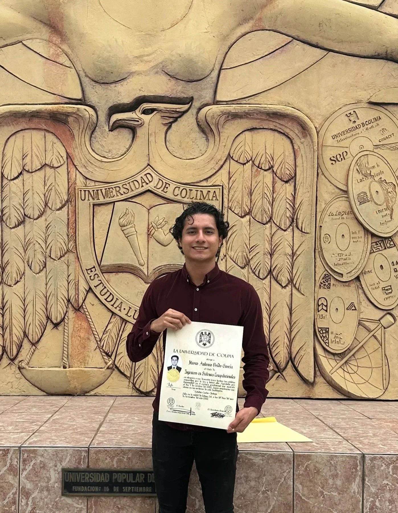 A young man standing in front of a large carved wall holding a diploma from Universidad de Colima, with a decorative crest and academic symbols.