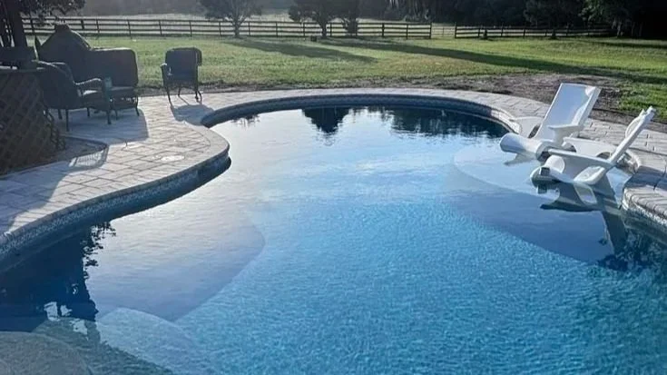 Swimming pool with lounge chairs on the pool deck, surrounded by a grassy yard and a fence in the background.