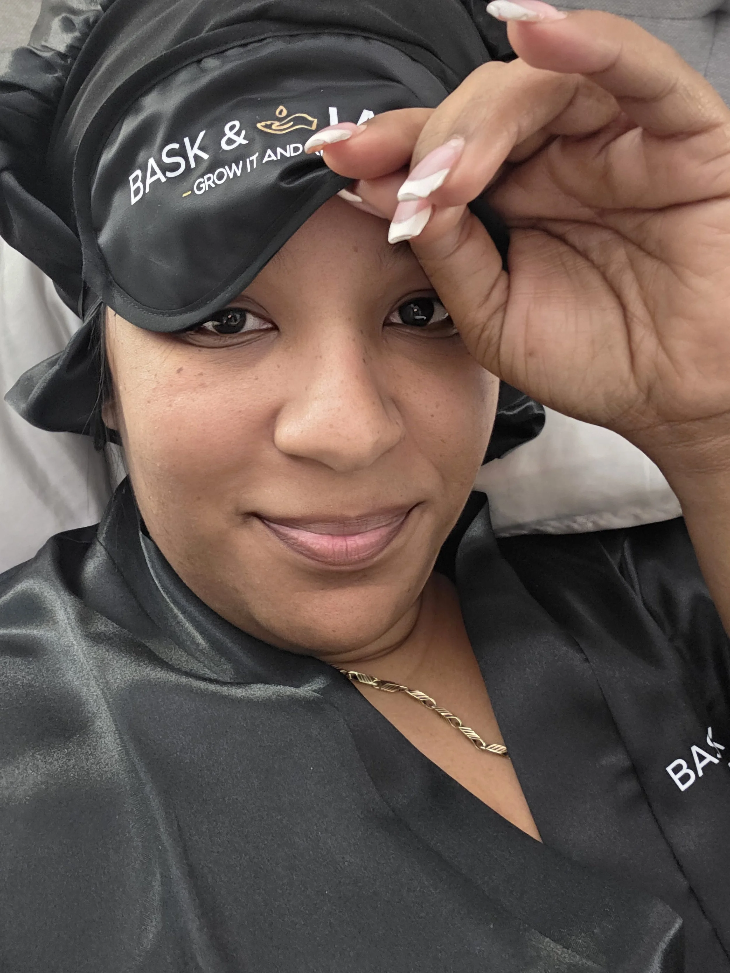 Gracefully Unfiltered author Tracey Joseph smiling while lying on a bed wearing Bask and Lather black satin hair cap and matching satin robe representing intentional rest and softness