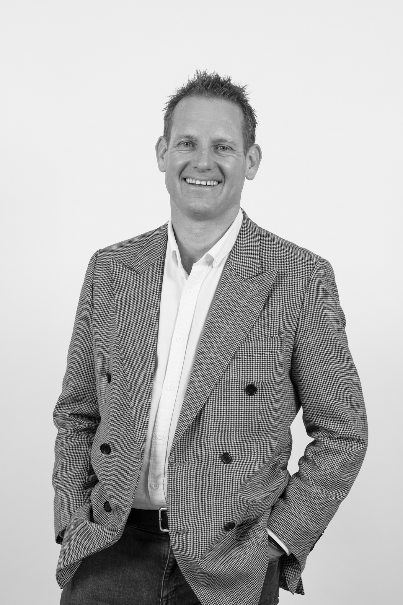 Black and white portrait of a smiling man wearing a checked blazer and white shirt, standing against a plain white background.