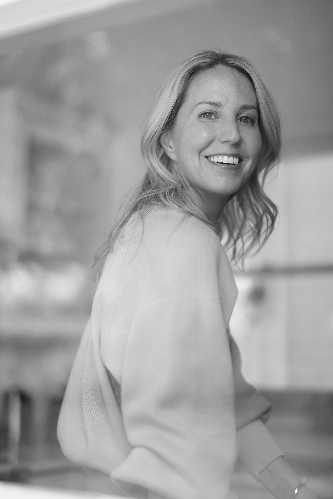 A smiling woman with wavy blonde hair wearing a light-colored sweater, sitting indoors with a blurred background.