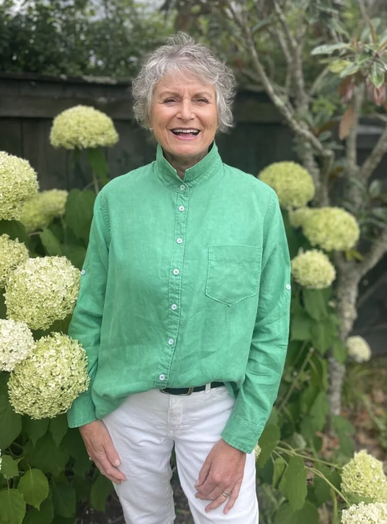 An elderly woman with gray curly hair smiling, wearing a green button-up shirt and white pants, standing outdoors in a garden with white hydrangea flowers and green foliage.