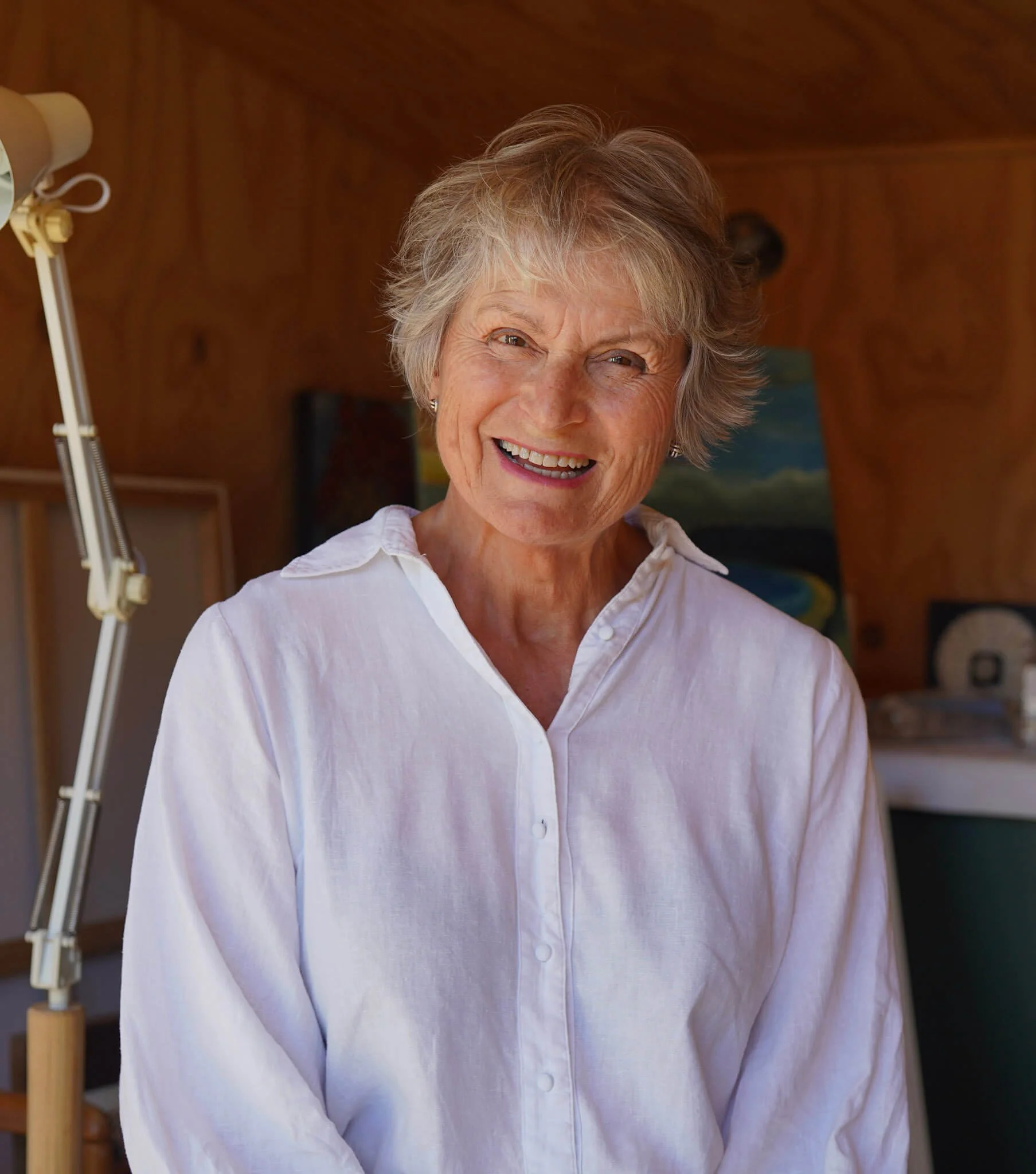A smiling elderly woman with short gray hair wearing a white button-up shirt, standing indoors with wooden walls and a painting in the background.
