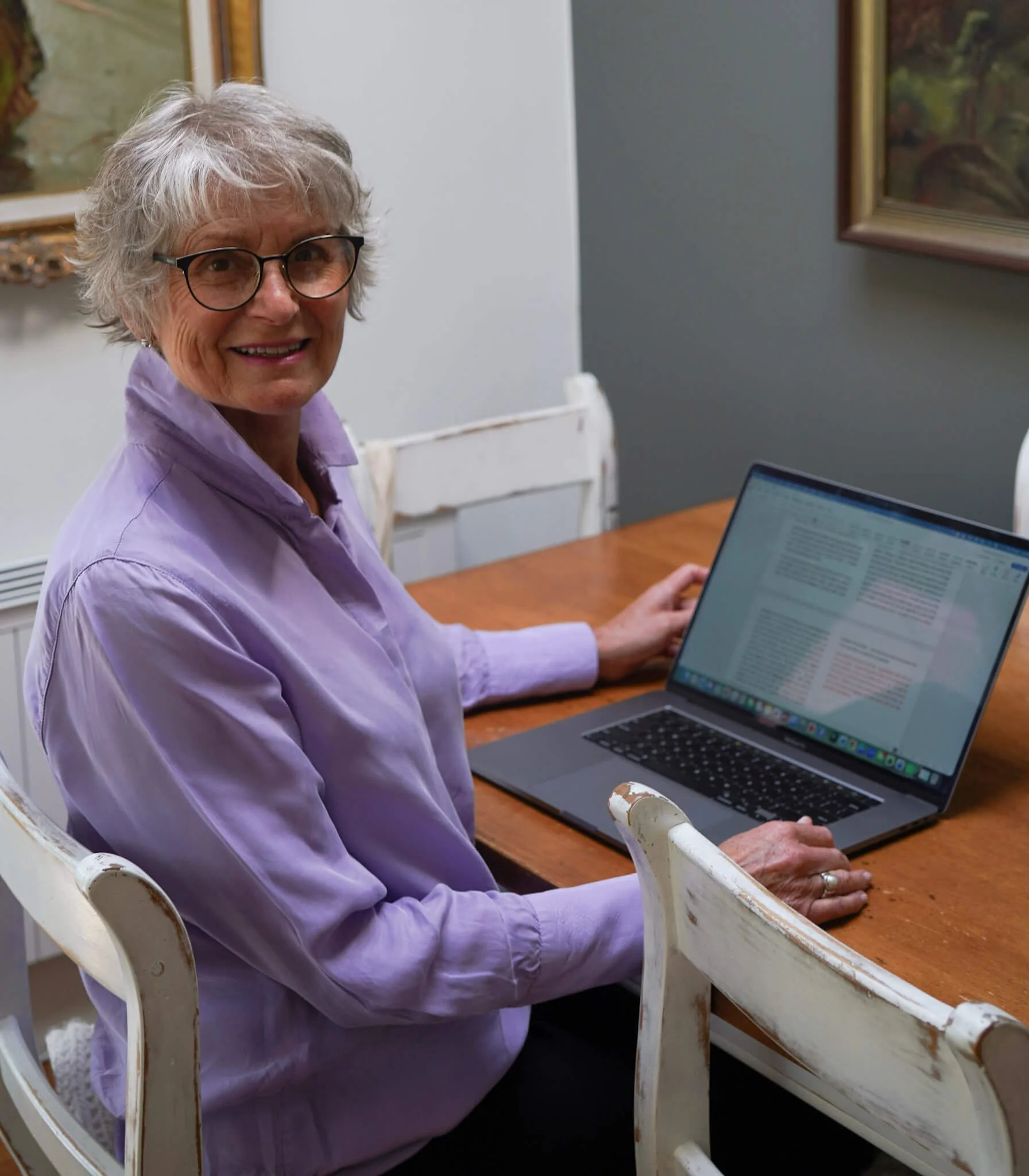 An elderly woman with gray hair and glasses, wearing a purple blouse, sitting at a wooden table with a laptop, in a room with framed paintings on the wall.