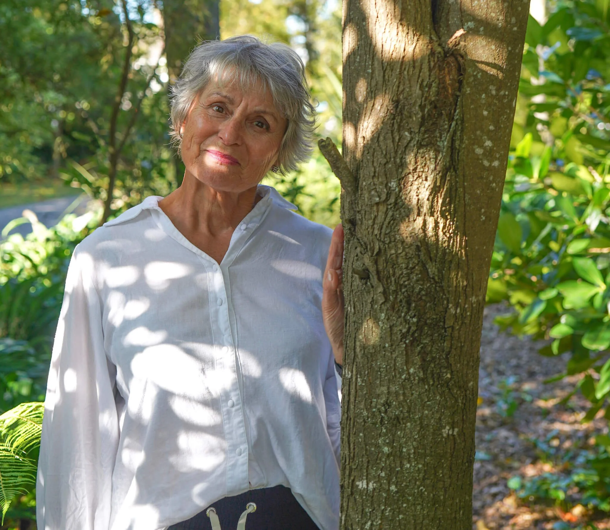 An elderly woman with short gray hair smiling softly while holding a tree trunk in a lush green park during daytime.
