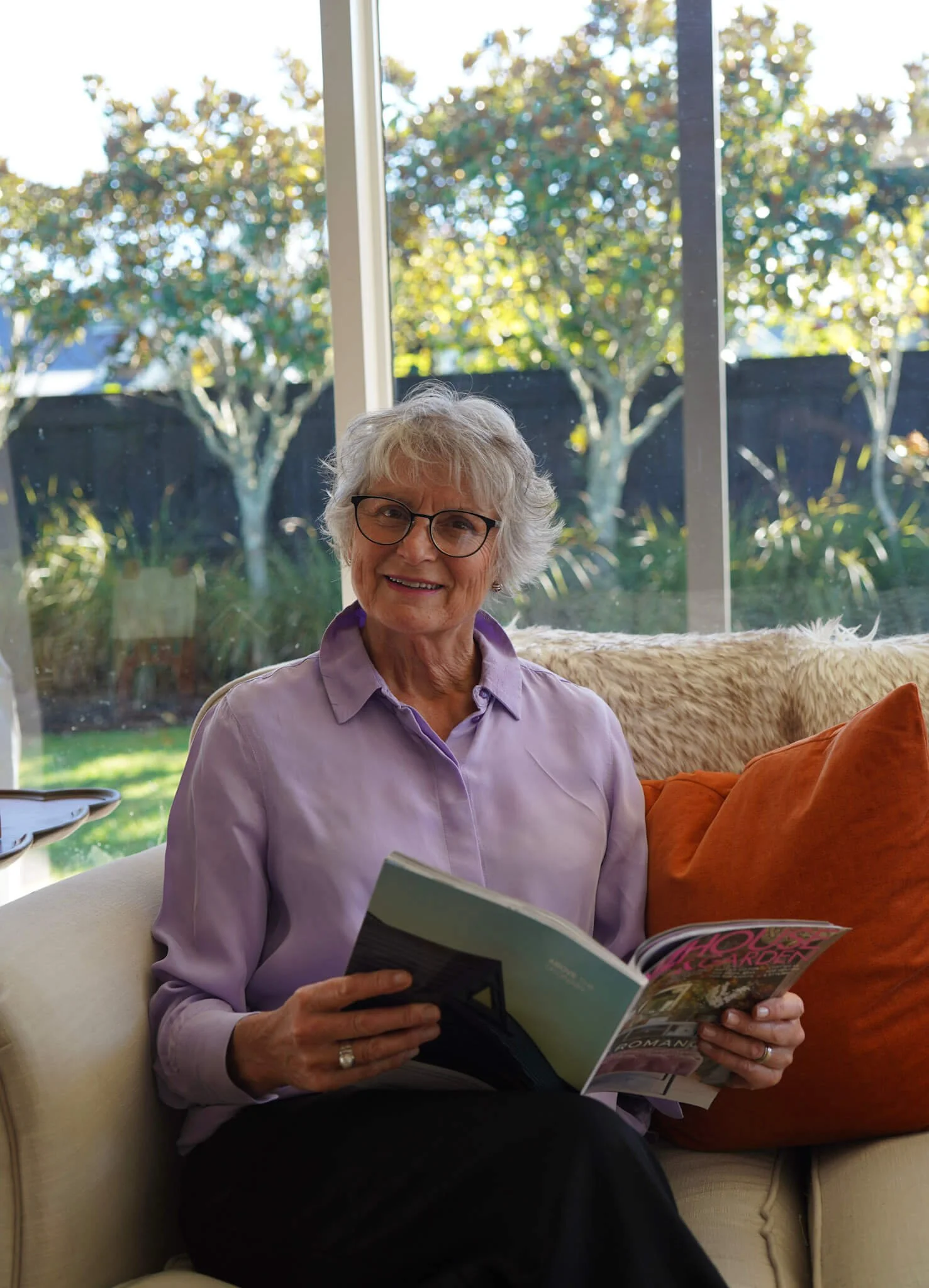 An elderly woman with gray hair, glasses, and a purple blouse sitting on a cream-colored sofa, reading a magazine in a bright room with large windows and a view of green trees outside.