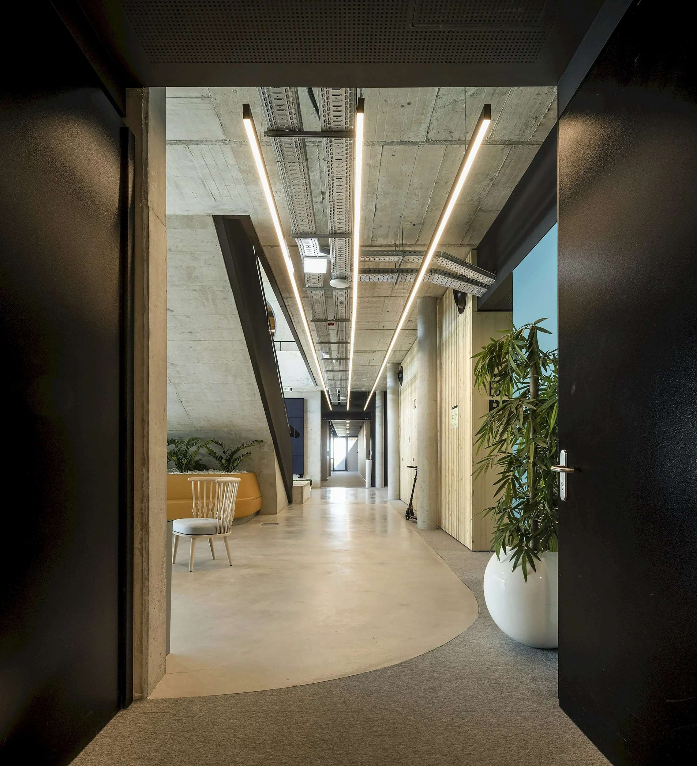 Modern office hallway with concrete ceilings, linear ceiling lights, and a mix of wood and black walls, featuring plants and a scooter.