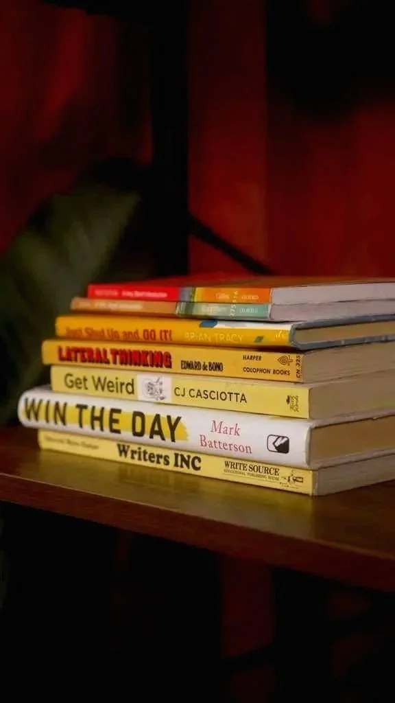 Stack of books on a wooden table in a dimly lit room, some titles include 'LITERAL THINKING,' 'Get Weird,' 'WIN THE DAY,' and 'Write Source.' A person wearing green clothing is partially visible in the background.