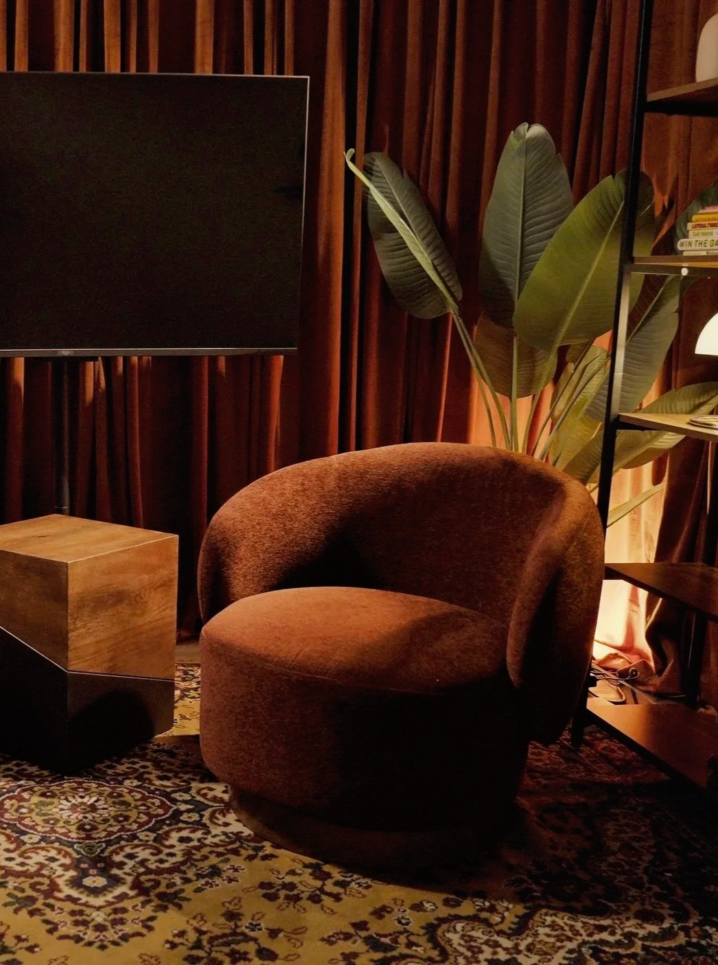 A cozy room corner with a plush brown velvet armchair, a small wooden side table, a large plant with broad green leaves, a patterned carpet, and a wall-mounted flat-screen TV. There are tall, rust-colored curtains and a black metal bookshelf with books.