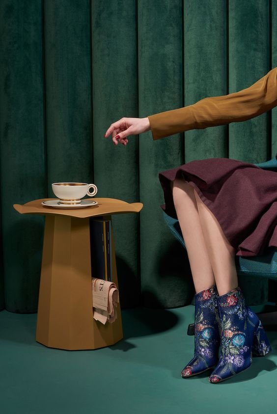 A person sitting on a teal chair with crossed legs, wearing a maroon skirt and embroidered ankle boots, reaching hand towards a cup on a small yellow table in front of a green curtain.