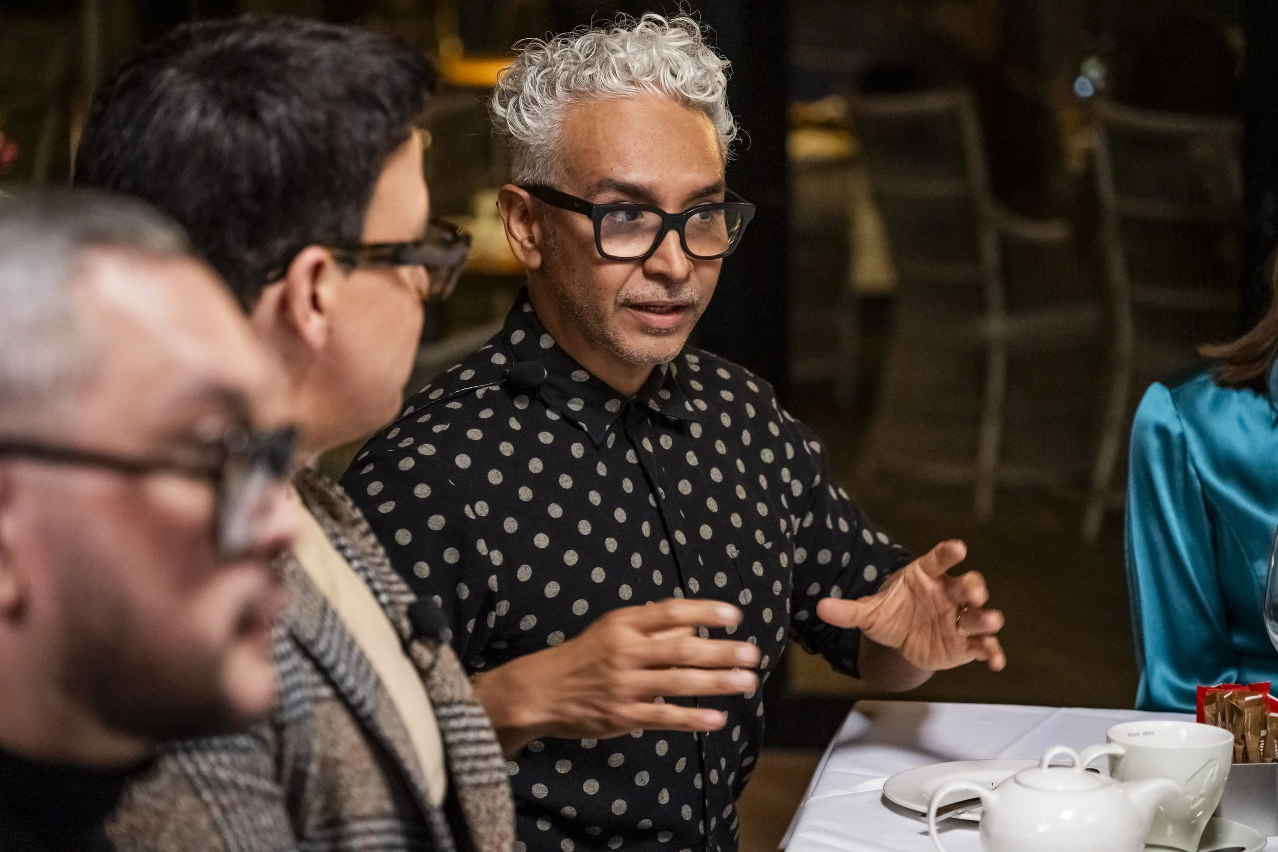 A man with gray curly hair and large black glasses, wearing a black shirt with white polka dots, speaking and gesturing with his hands at a table during a discussion with other people.