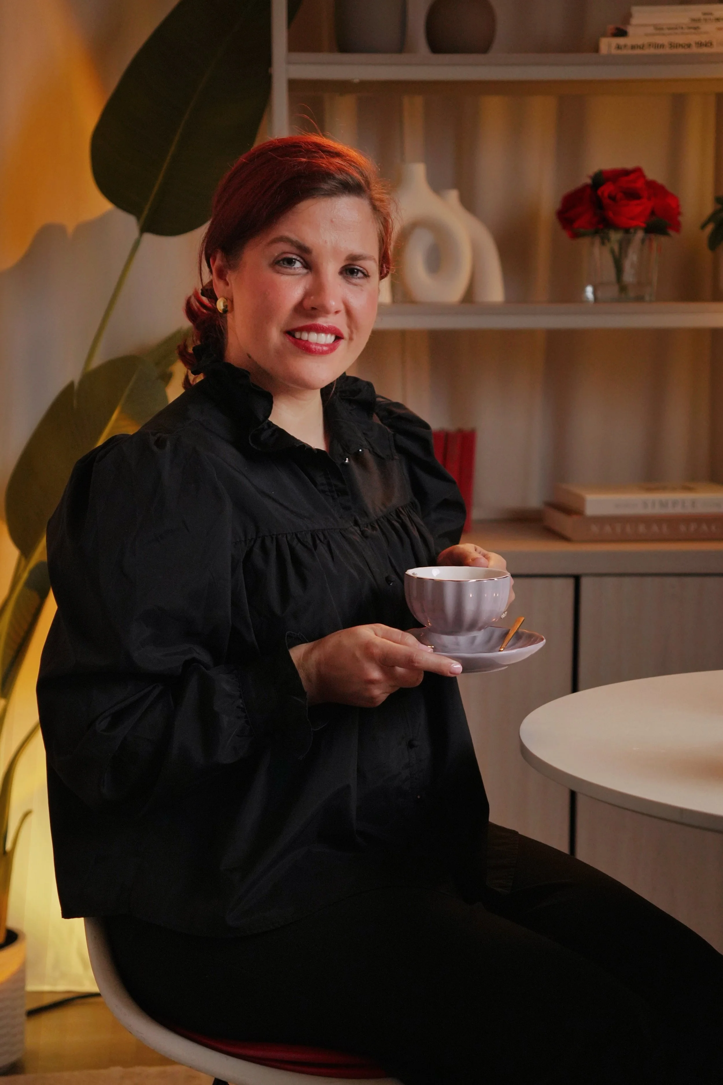 Woman with red hair, wearing a black blouse, holding a white cup and saucer, sitting in a room with decorative shelves and plants.