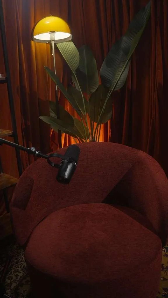 A red velvet chair, a yellow floor lamp, large green leaves of a potted plant, and a wooden wall with curtains in a cozy room.