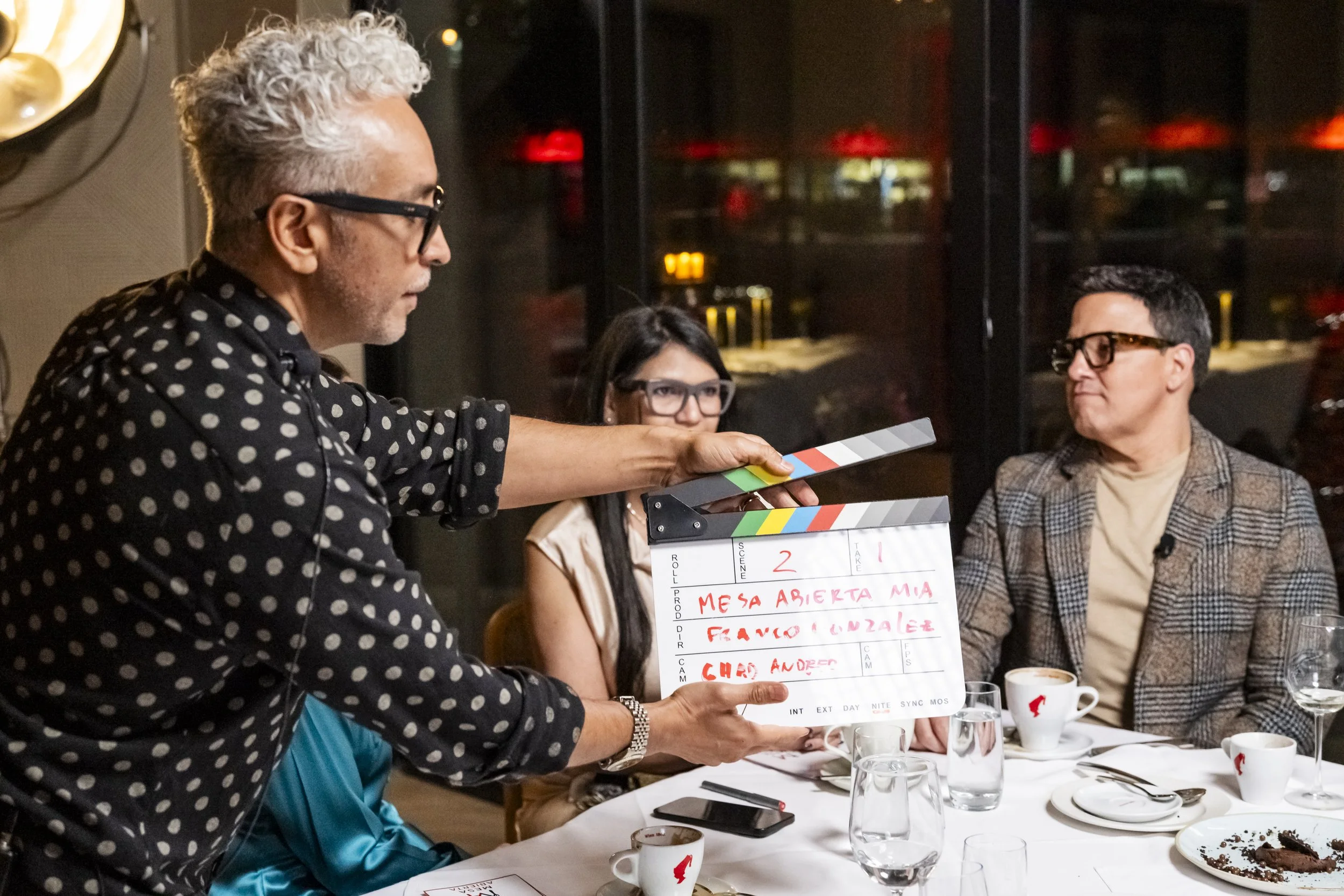 Filmmaker holding a clapperboard at a dinner table with three people, two women and one man, all wearing glasses, in a restaurant setting.