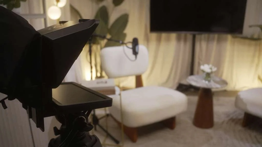 Interior studio setup with film lighting, two white armchairs, a small round wooden table with a flower vase, a large wall-mounted TV, and a yellow curtain backdrop.