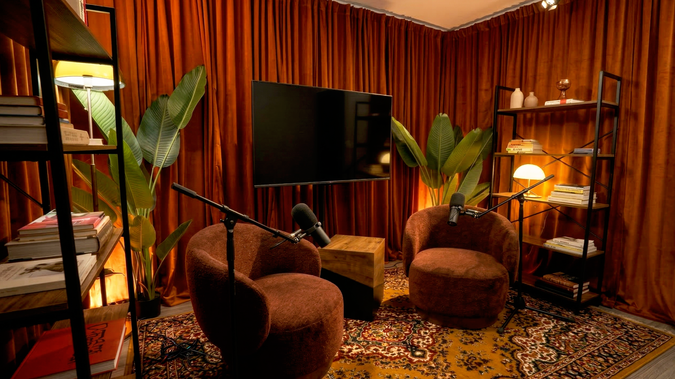 Cozy living room with orange curtains, large green potted plants, two rust-colored velvet chairs with microphones, a flat-screen TV mounted on the wall, wooden side table, two black metal bookshelves with books, decorative vases, table lamp, patterne