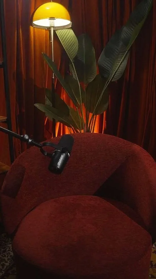 A red velvet armchair with a black microphone resting on it, a tall floor lamp with a yellow shade, and large green leaves in the background, all set against wooden paneling.