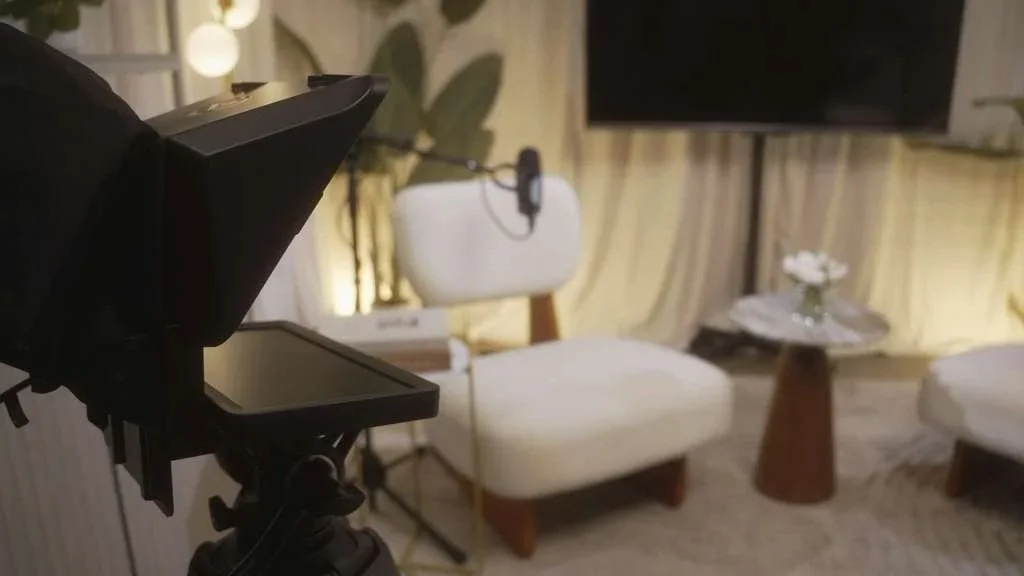 Camera filming a cozy TV living room with two white armchairs, a small round table with a flower vase, a wall-mounted TV, and curtains.