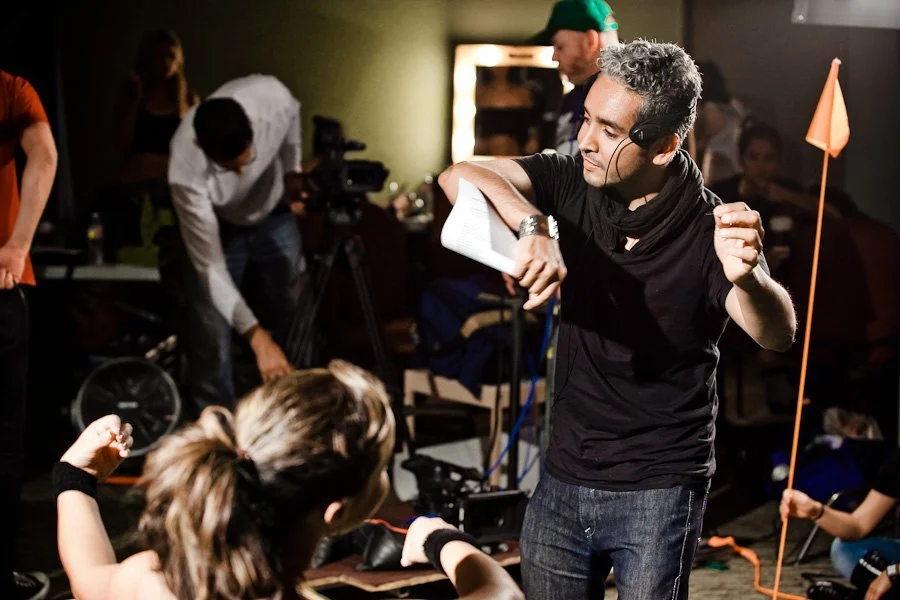 A film director giving instructions to a woman on set during a production. The director is wearing a headset, sunglasses, and a black shirt, and is holding a script. In the background, crew members and filming equipment are visible.