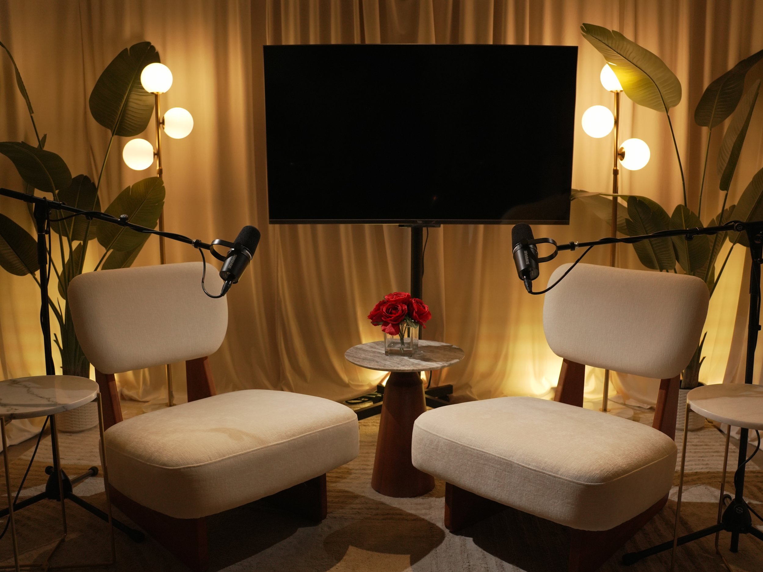 Two beige chairs with microphones, a small round marble table with red roses, a large TV, and plants with lights in a cozy room.