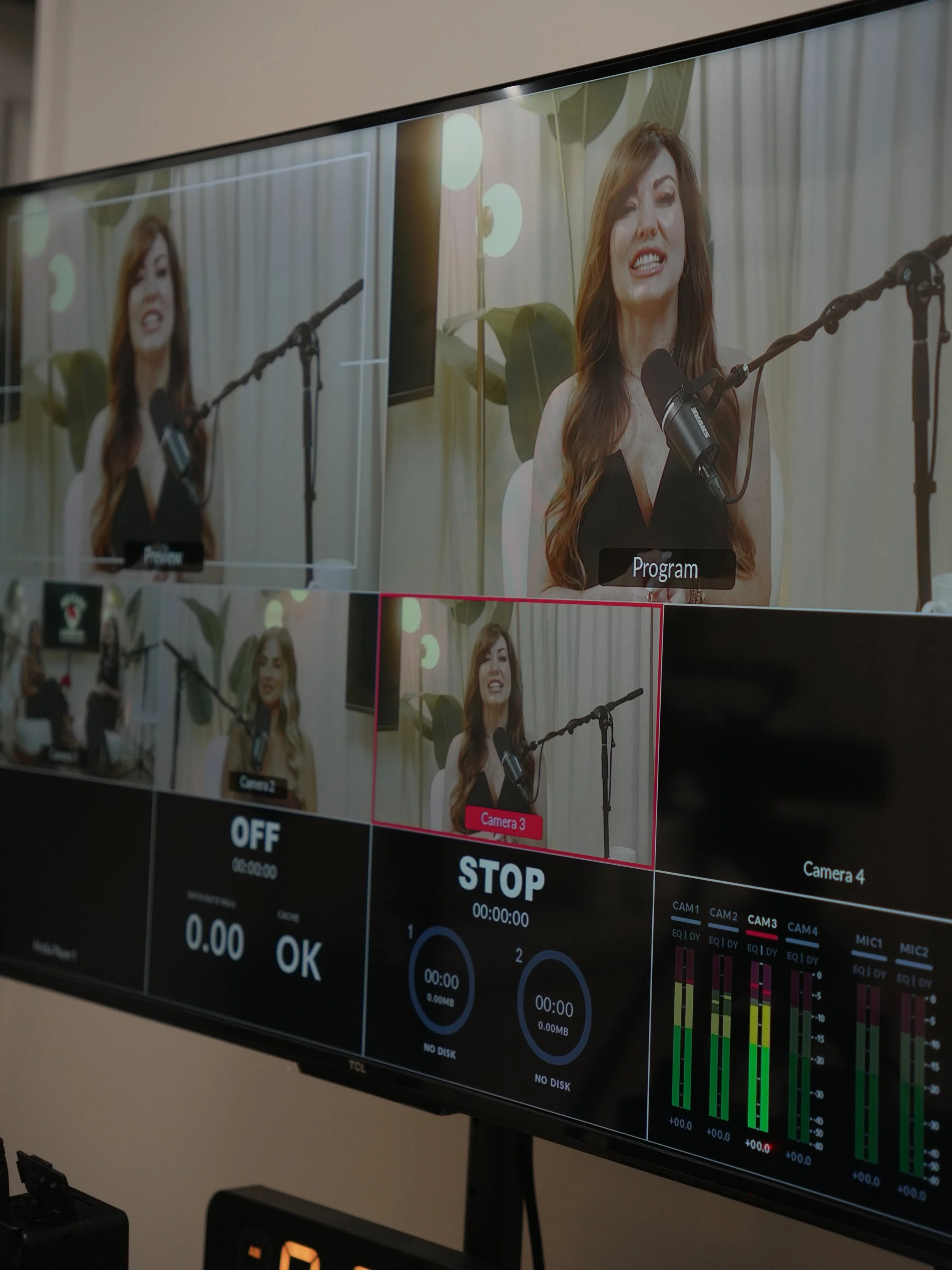 A professional video monitor displaying a woman speaking into a microphone during a recording session, with multiple camera angles and audio levels shown on the screen.