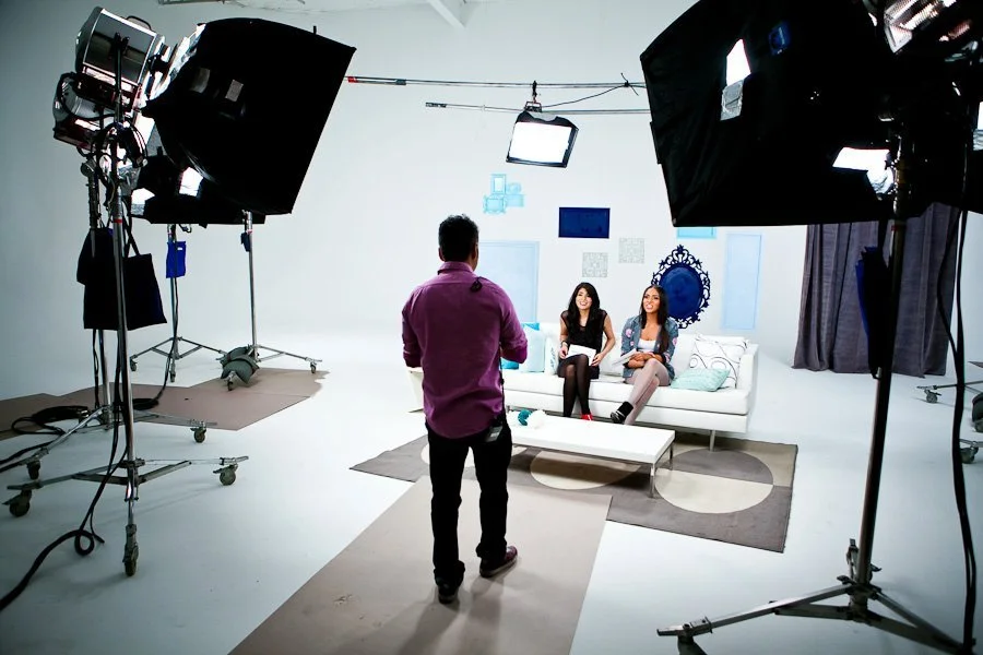 Two women sitting on a white sofa in a TV studio, being filmed by a man standing in front of them, with professional lighting and camera equipment around.