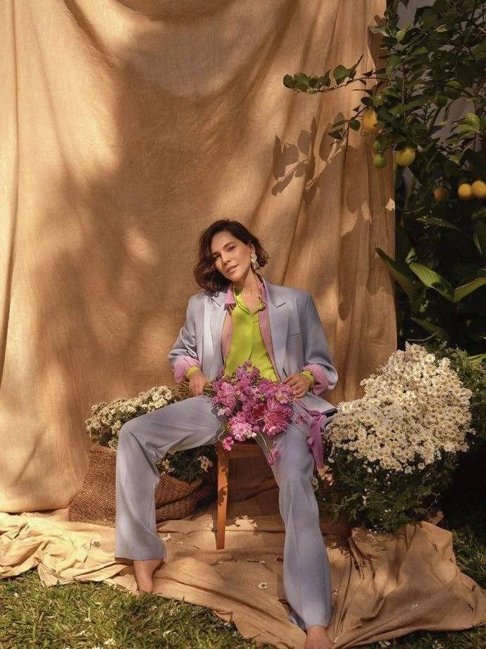 Woman sitting on a chair surrounded by flowers, wearing a pastel suit with a lime green shirt, set against a beige backdrop with greenery and flowers.