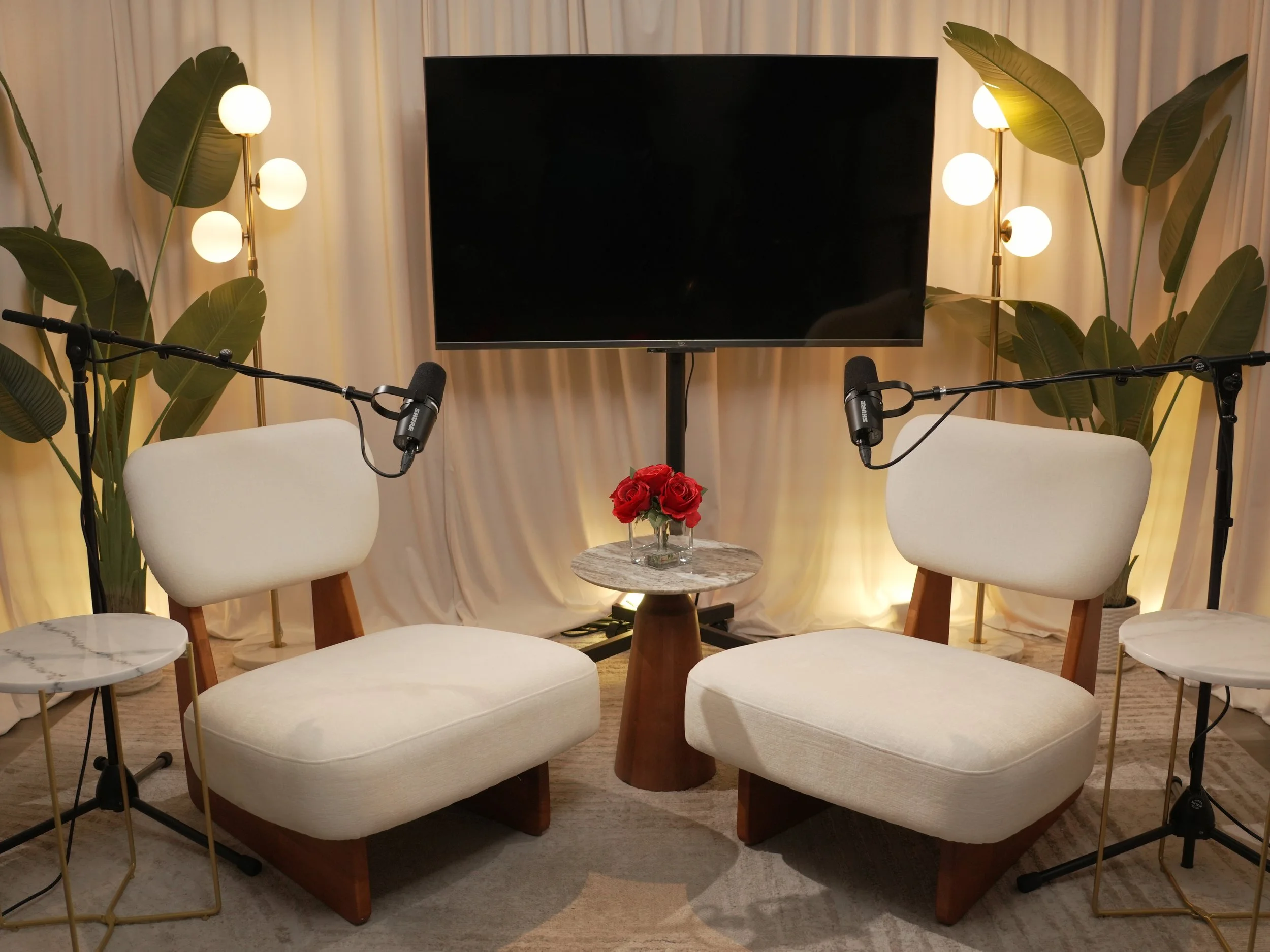 A cozy podcast studio setup with two white upholstered chairs facing each other, each equipped with a microphone on a stand. Between the chairs is a small round side table with a vase of red roses. Behind is a large flat-screen TV mounted on the wall