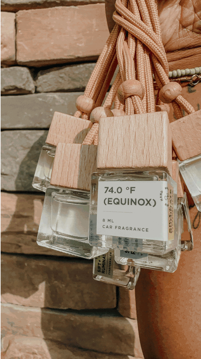 A bundle of car fragrance diffusers with wooden caps and light brown rope handles, labeled '74.0°F Equinox,' hanging against a stone wall.