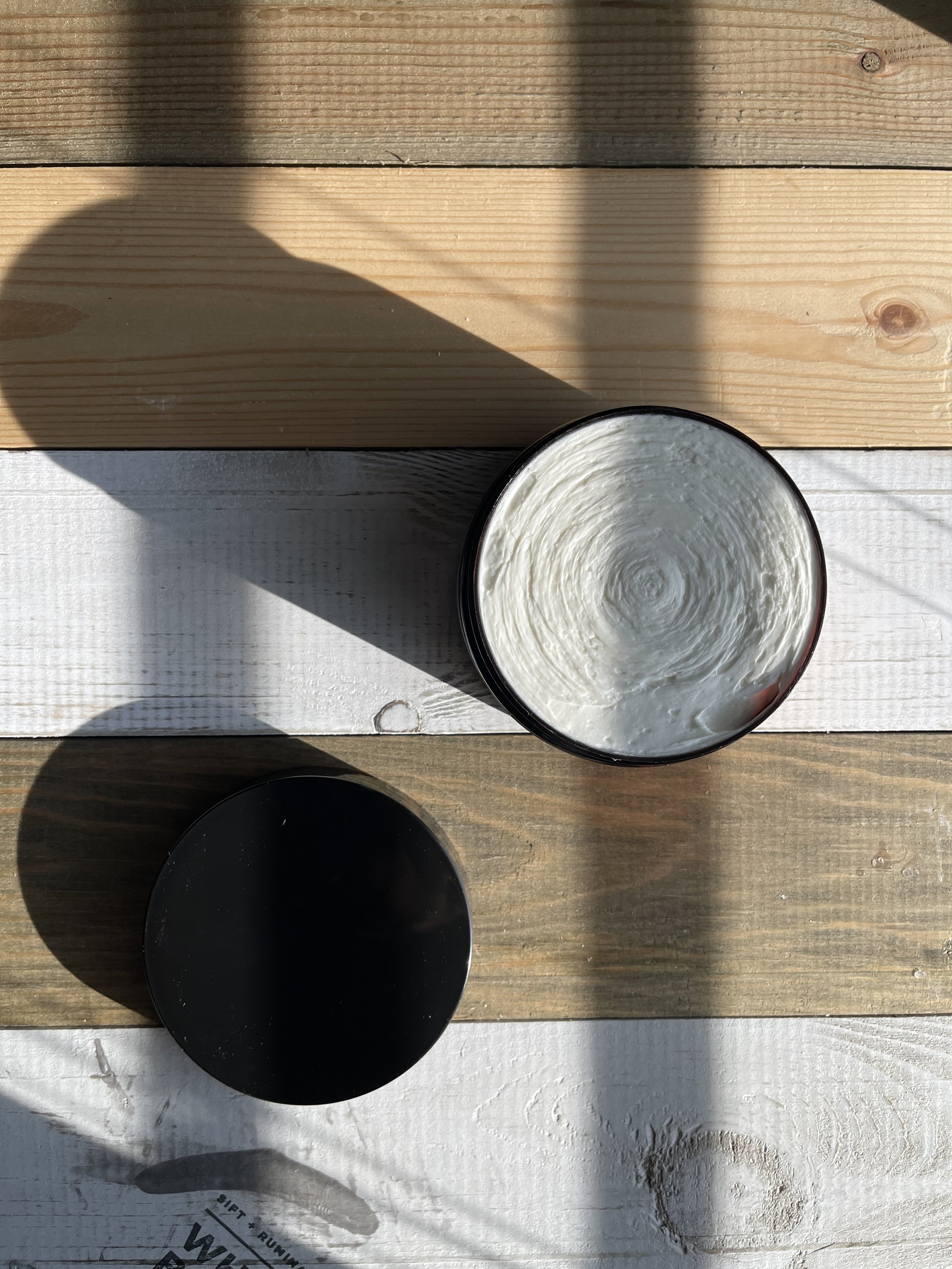 A black container filled with shea butter, placed on a wooden surface with a shadow cast over it.
