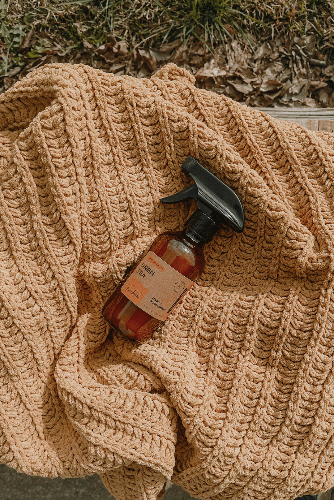 A spray bottle labeled 'Urban Tea' sitting on a plush, textured, peach-colored blanket outdoors with leaf-covered ground in the background.