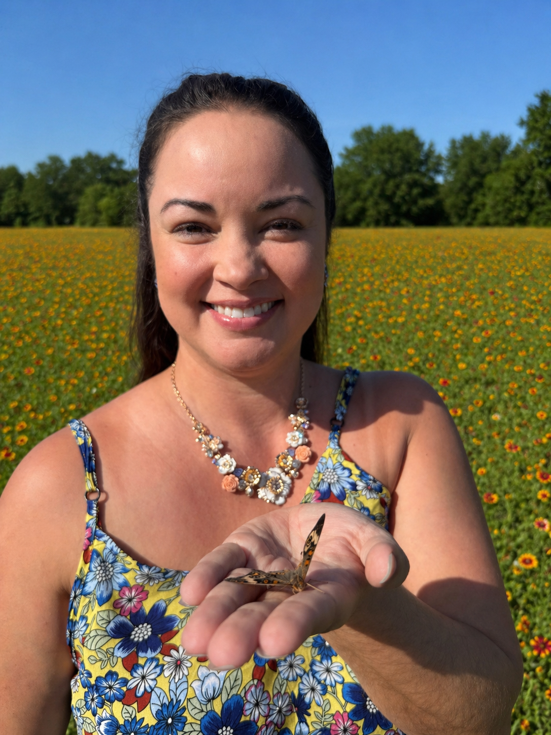 A woman smiling and holding a butterfly in her hand in a field of yellow and orange flowers with a blue sky and green trees in the background.