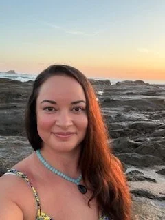 A woman with long brown hair wearing a colorful dress and a turquoise necklace, standing on a rocky beach at sunset.