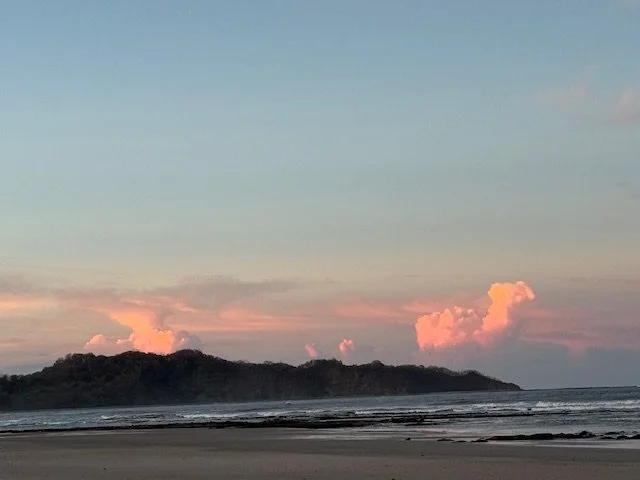 A peaceful beach scene during sunset with a cloudy sky and a small hill or island in the distance.