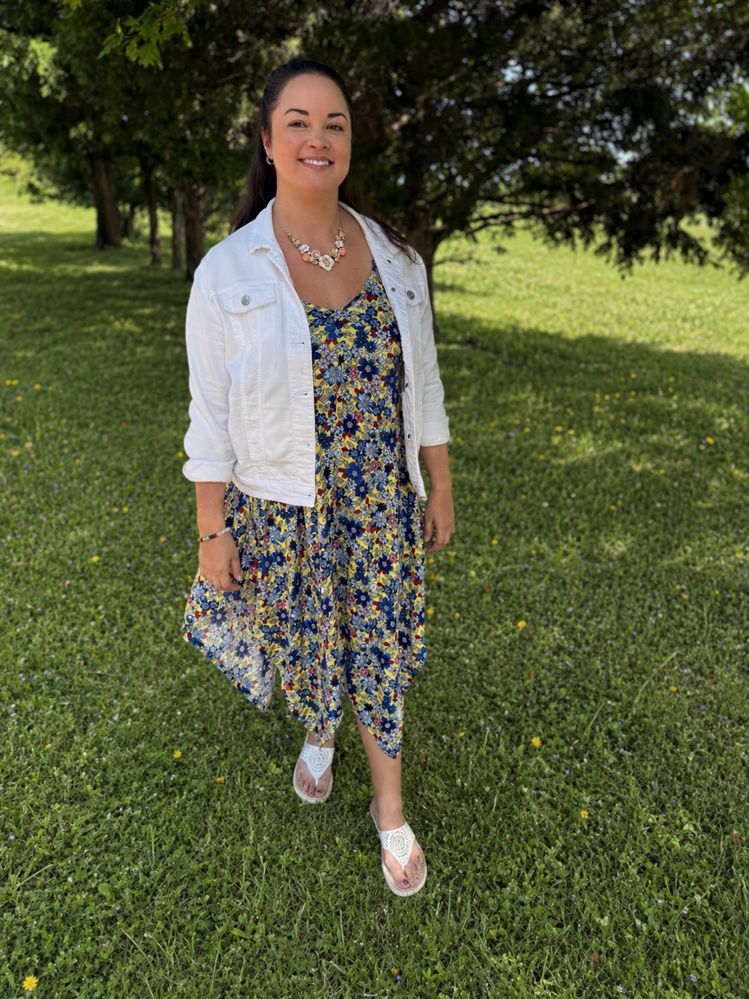 A woman standing outdoors on a grassy field with trees in the background, wearing a floral dress, white jacket, and white sandals, smiling.