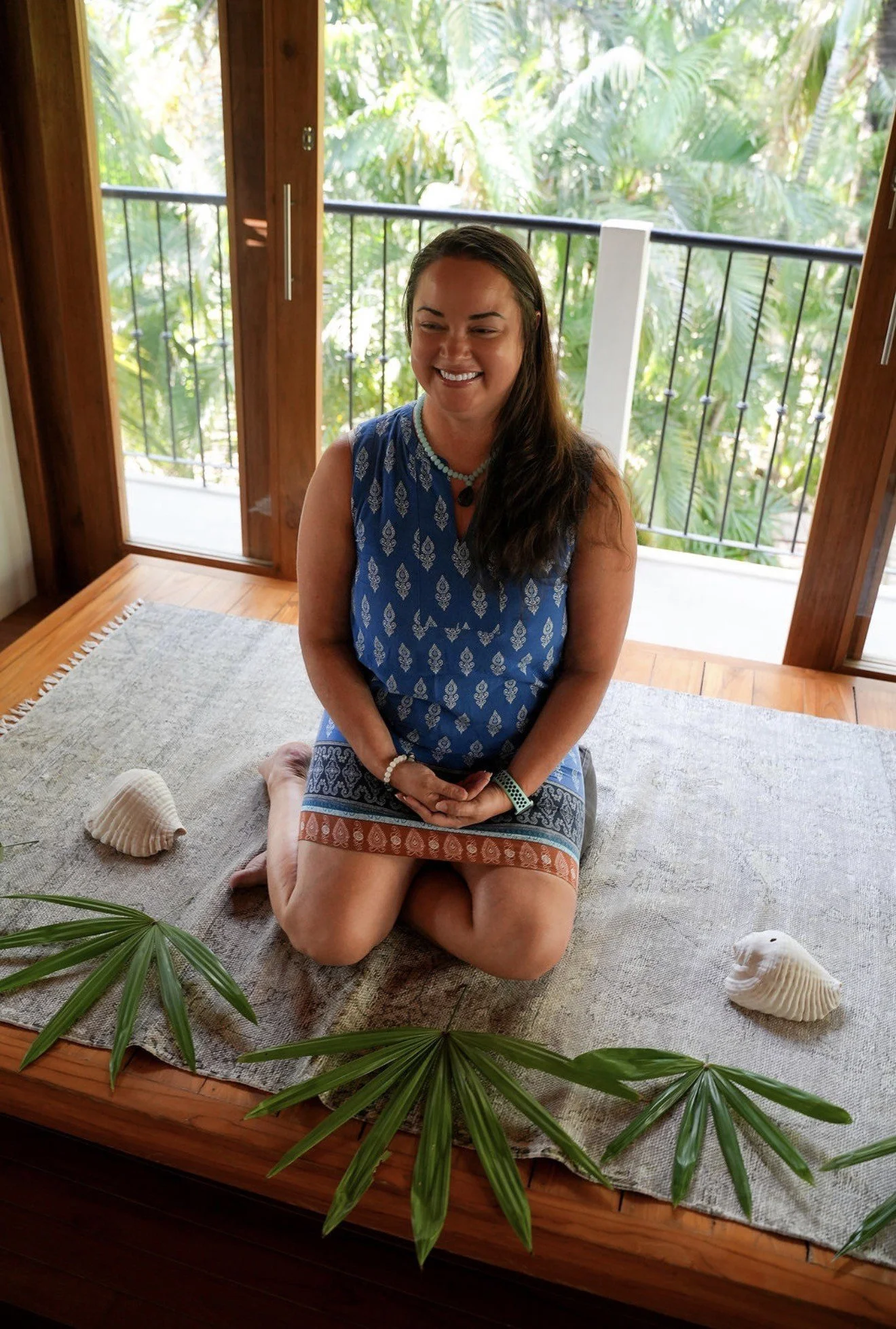 A woman kneels on a beige rug, smiling with eyes closed, in a room with large windows showing lush green trees outside. She wears a blue dress with white patterns, necklaces, and a bracelet. Seashells and green leaves decorate the rug.