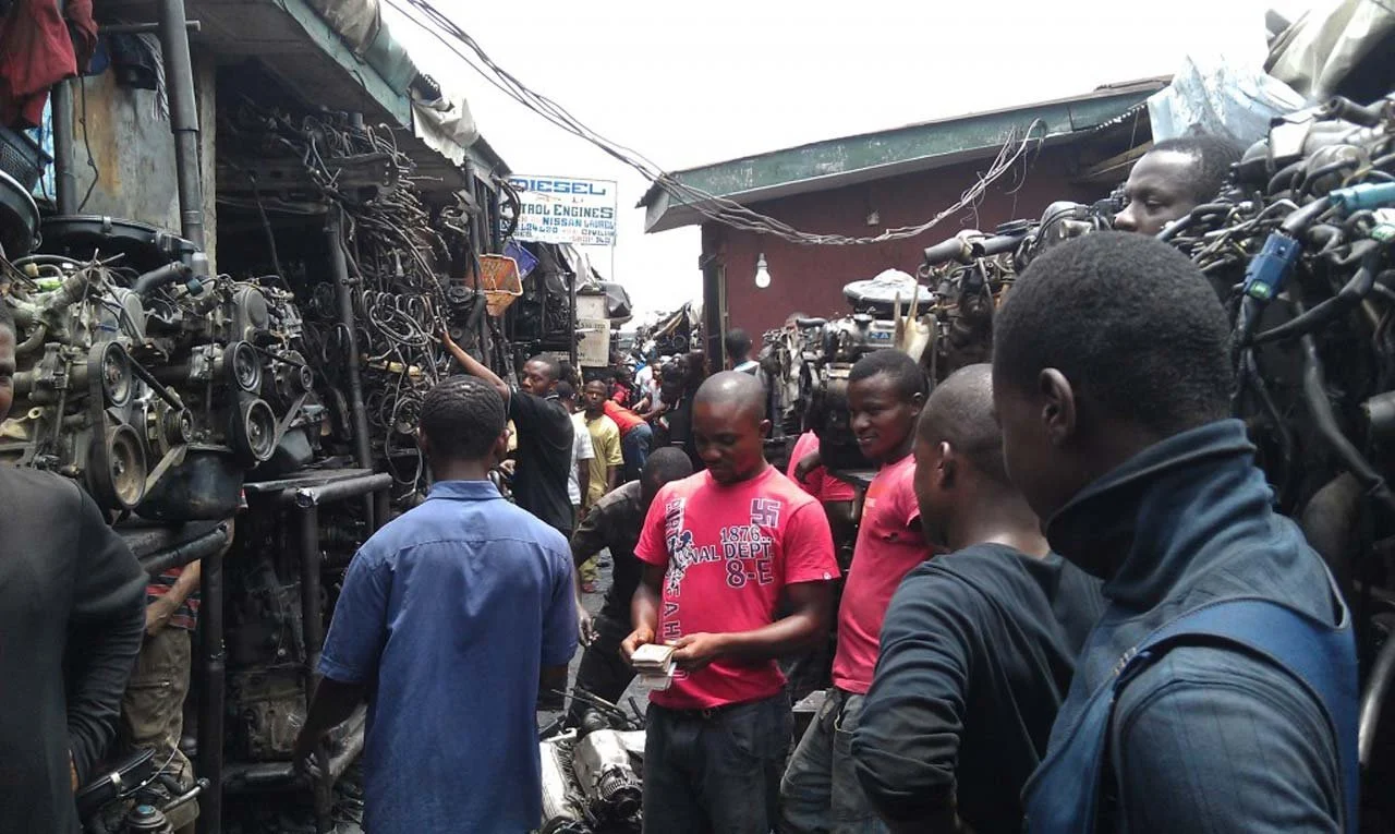 A crowded marketplace with people gathered around various car engines for sale, some inspecting and others chatting or holding money in an outdoor setting.
