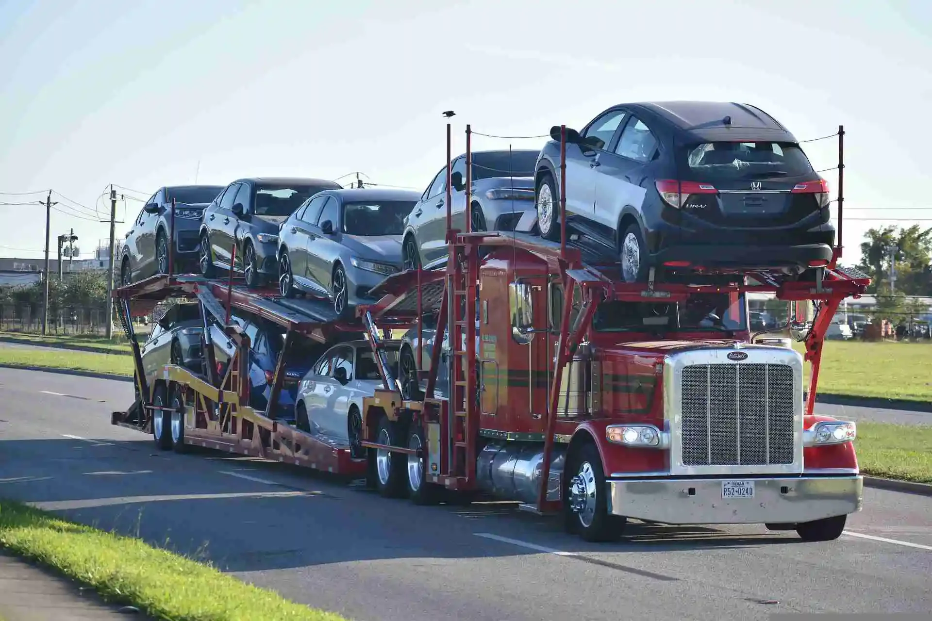 A red car carrier truck transporting multiple cars on a road with a grassy area and power lines in the background.