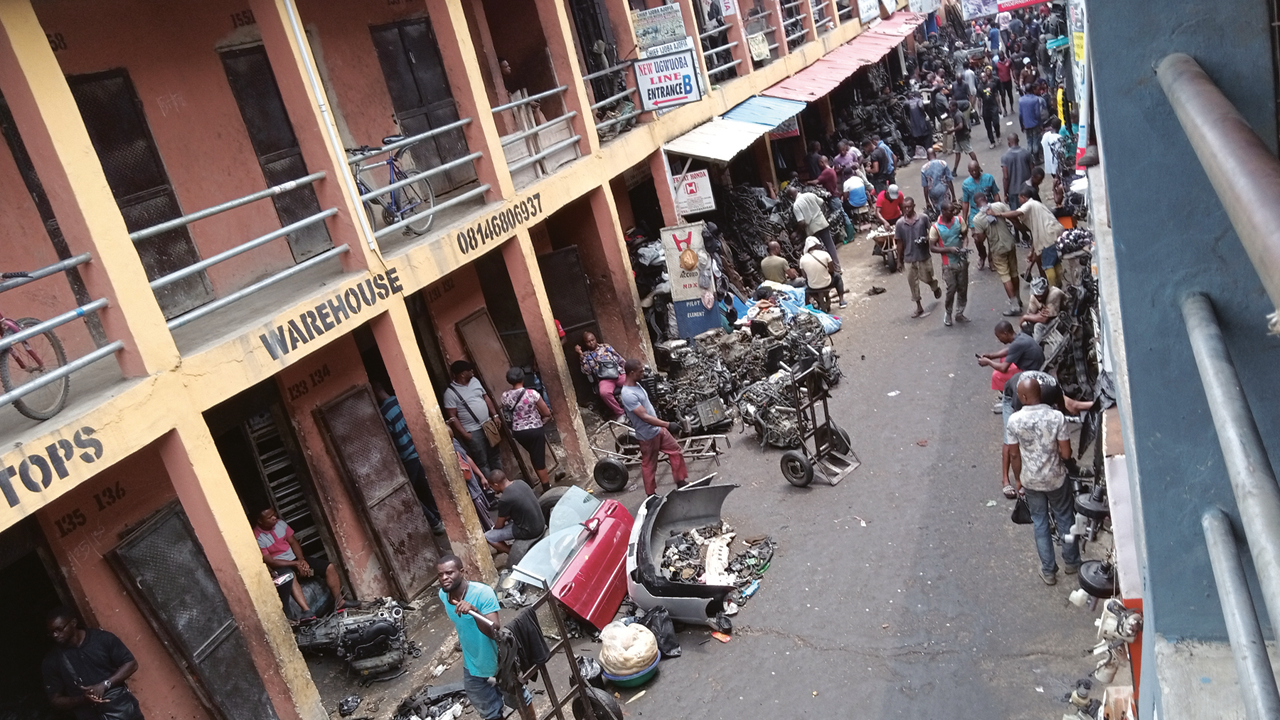 Street scene with vendors, motorcycle parts, and people walking in a crowded marketplace area adjacent to a building with balconies.