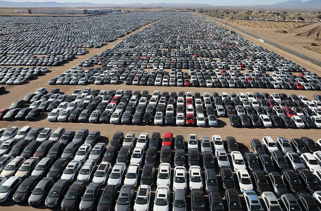 A vast car lot filled with numerous new vehicles parked in rows, with mountains visible in the background.