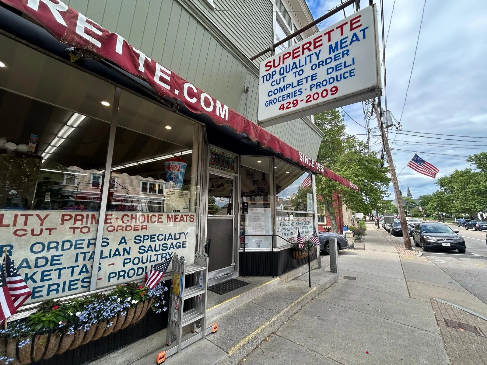 Exterior view of Superette, a grocery store with signs indicating fresh meats, Italian sausage, and poultry. The store has patriotic decorations with small American flags and a large American flag is visible on a pole. Cars are parked along the street, and there are trees and a cloudy sky in the background.