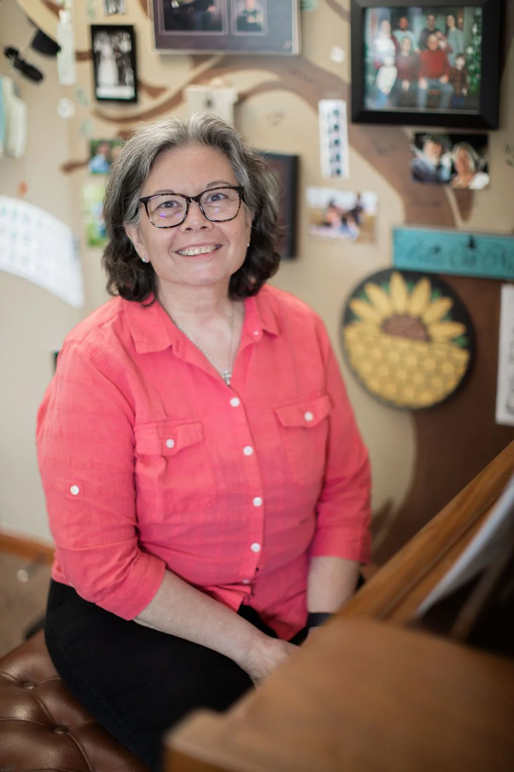 A woman with glasses and shoulder-length gray hair, wearing a coral pink button-up shirt, sitting at a piano in a cozy room decorated with family photos and artwork.