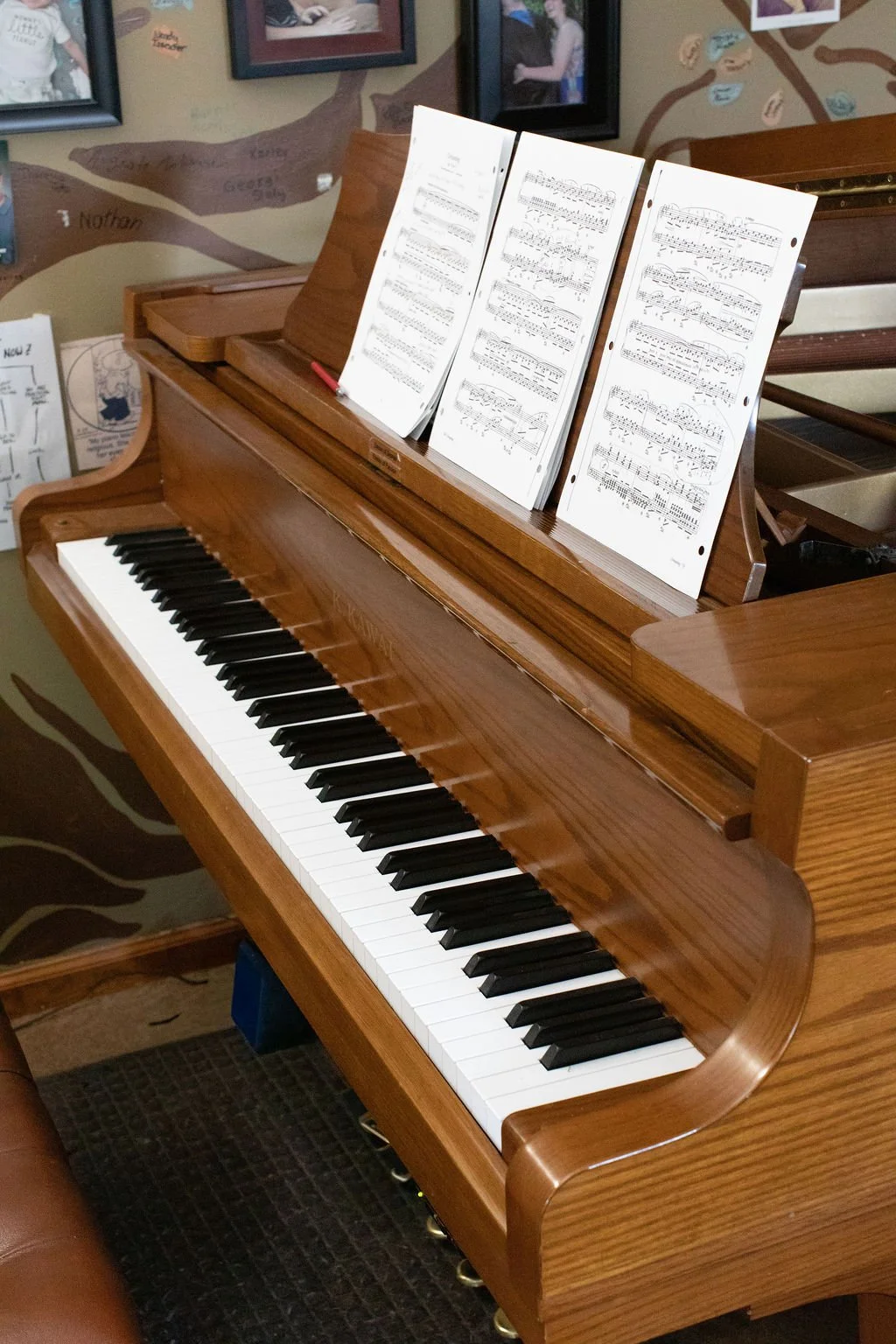 A wooden upright piano with black and white keys, sheet music on the music stand, and framed photographs on the wall behind.