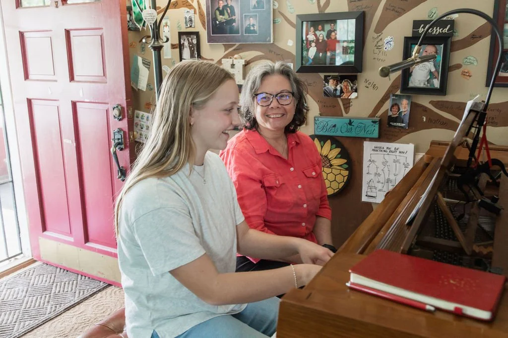 A young woman and an older woman are sitting together at a piano, smiling. The setting is a cozy, decorated room with framed photos and artwork on the wall behind them.