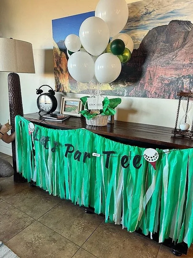 Decorated table with green and white paper fringe, birthday banner reading 'Lets Part Tee', and a bunch of white and green balloons, set in a room with a landscape painting and a lighthouse-shaped clock.
