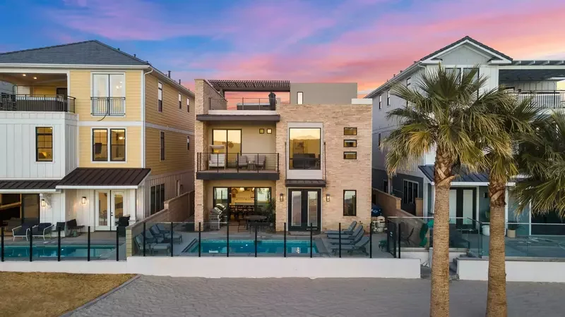 Modern three-story house with large windows, outdoor balconies, a backyard with a swimming pool, and palm trees, at sunset.