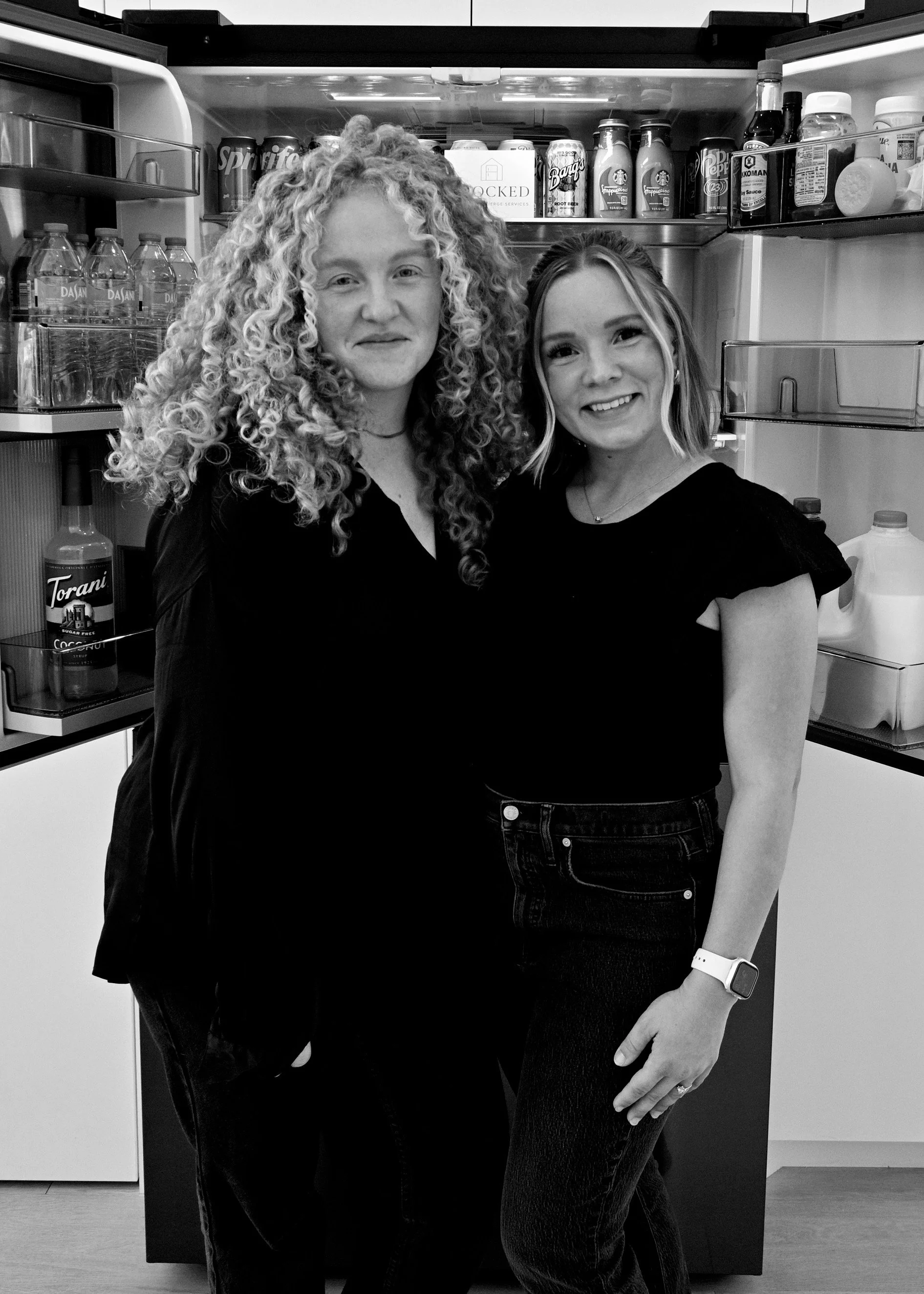 Two women smiling inside a refrigerator with beverages and condiments, one with curly hair, the other with straight hair.