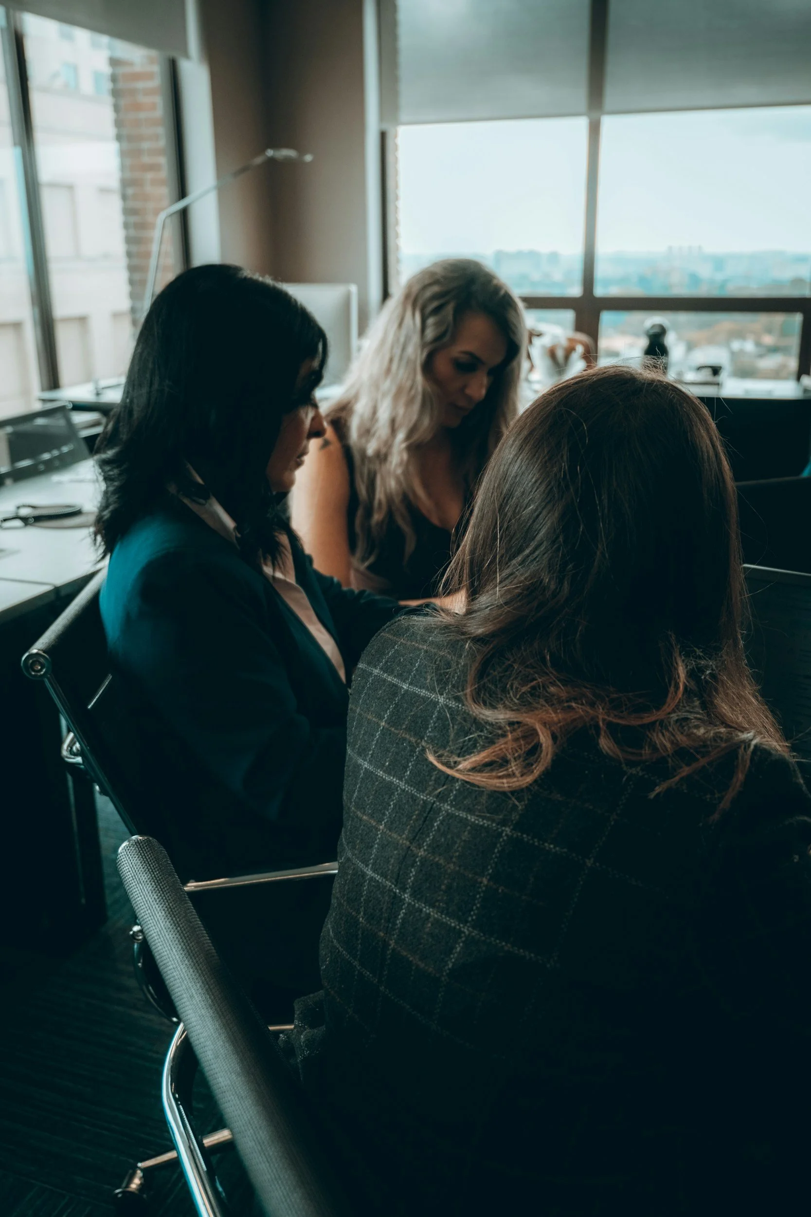 Three women sitting at a table in an office, engaged in a discussion, with large windows overlooking a cityscape in the background.