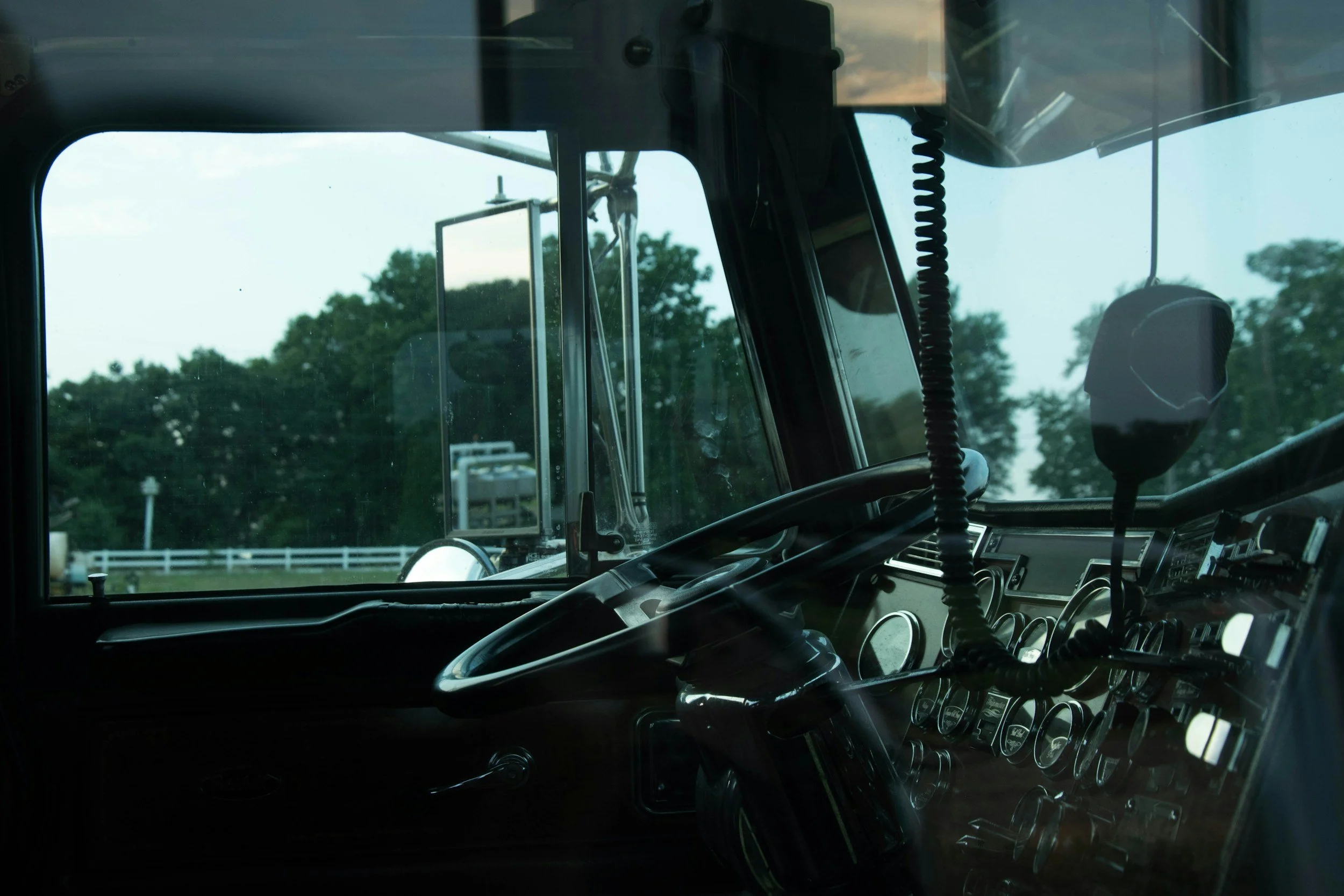 Inside a vehicle cabin with various gauges and controls, and a large window showing trees and a fence outside.