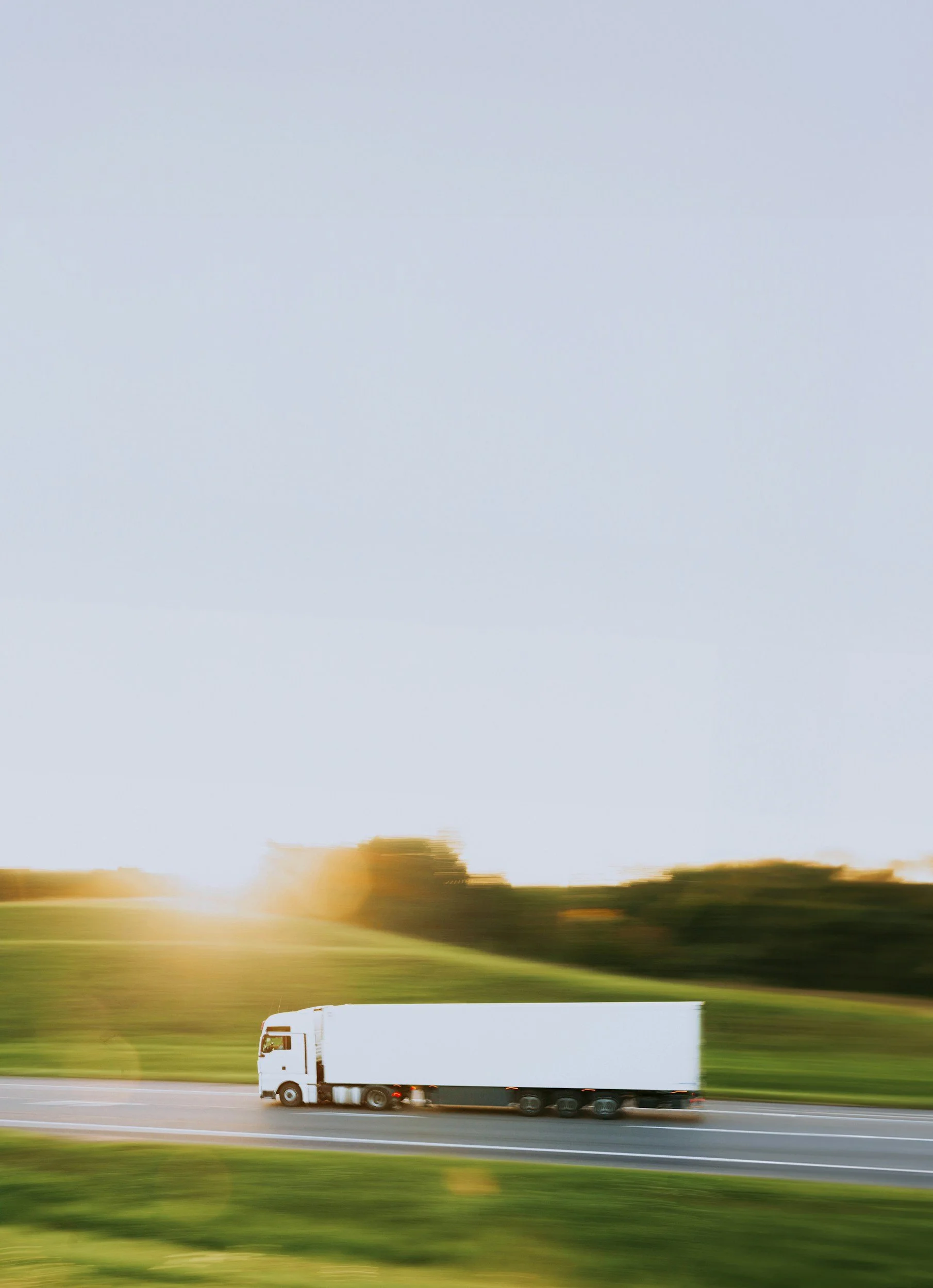 A white semi-truck driving on a highway with a blurred background of trees and sky during sunset.